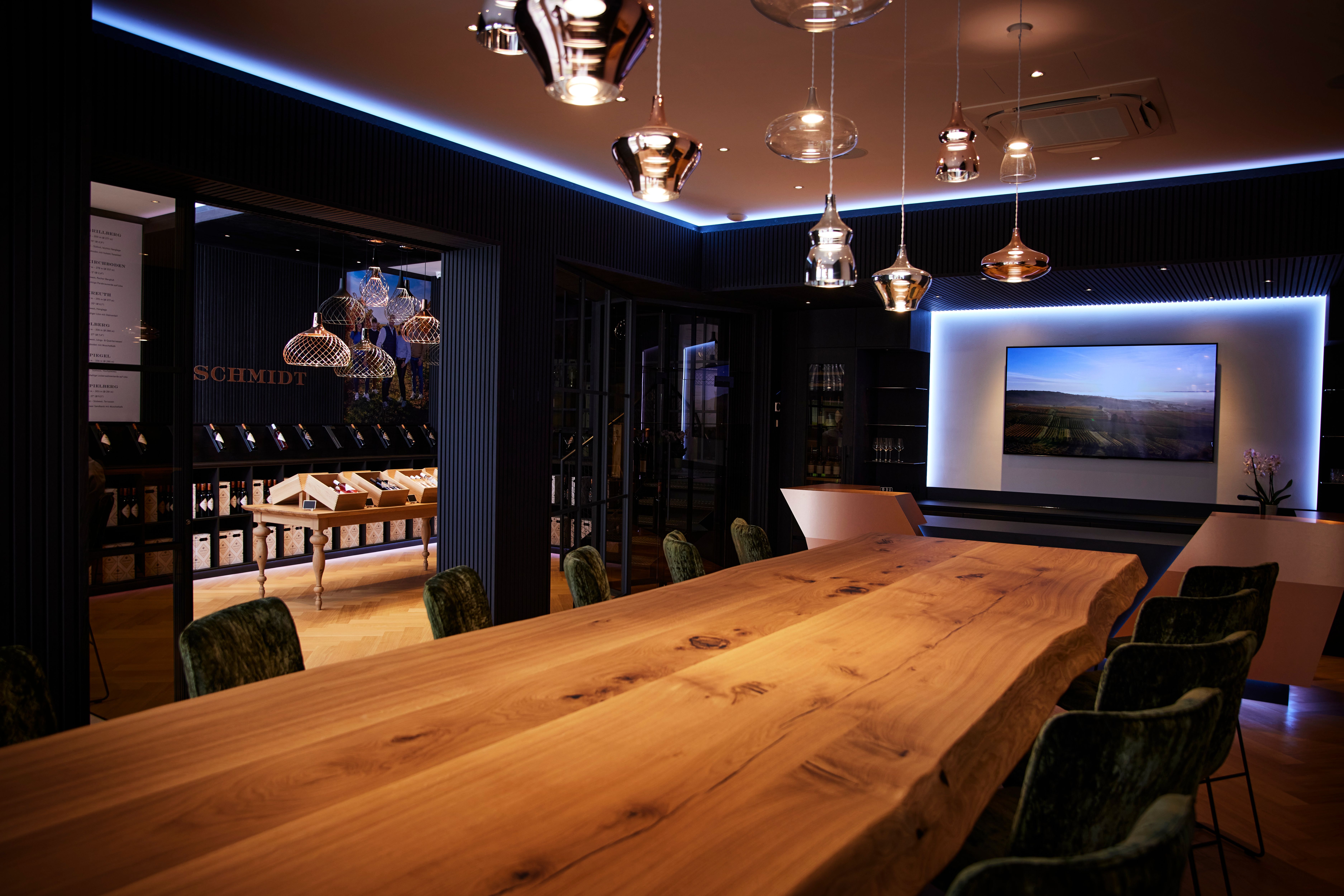 Eleganter Raum mit langem Holztisch, modernen Lampen und einem Bildschirm an der Wand.