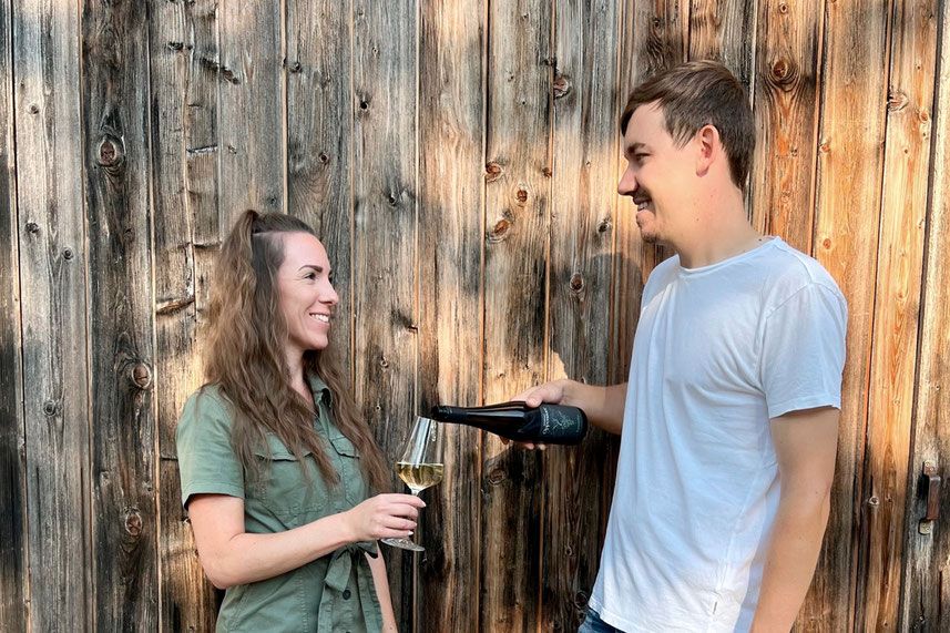 Ein Mann schenkt einer Frau Wein ein, beide lächeln vor einer Holzwand.