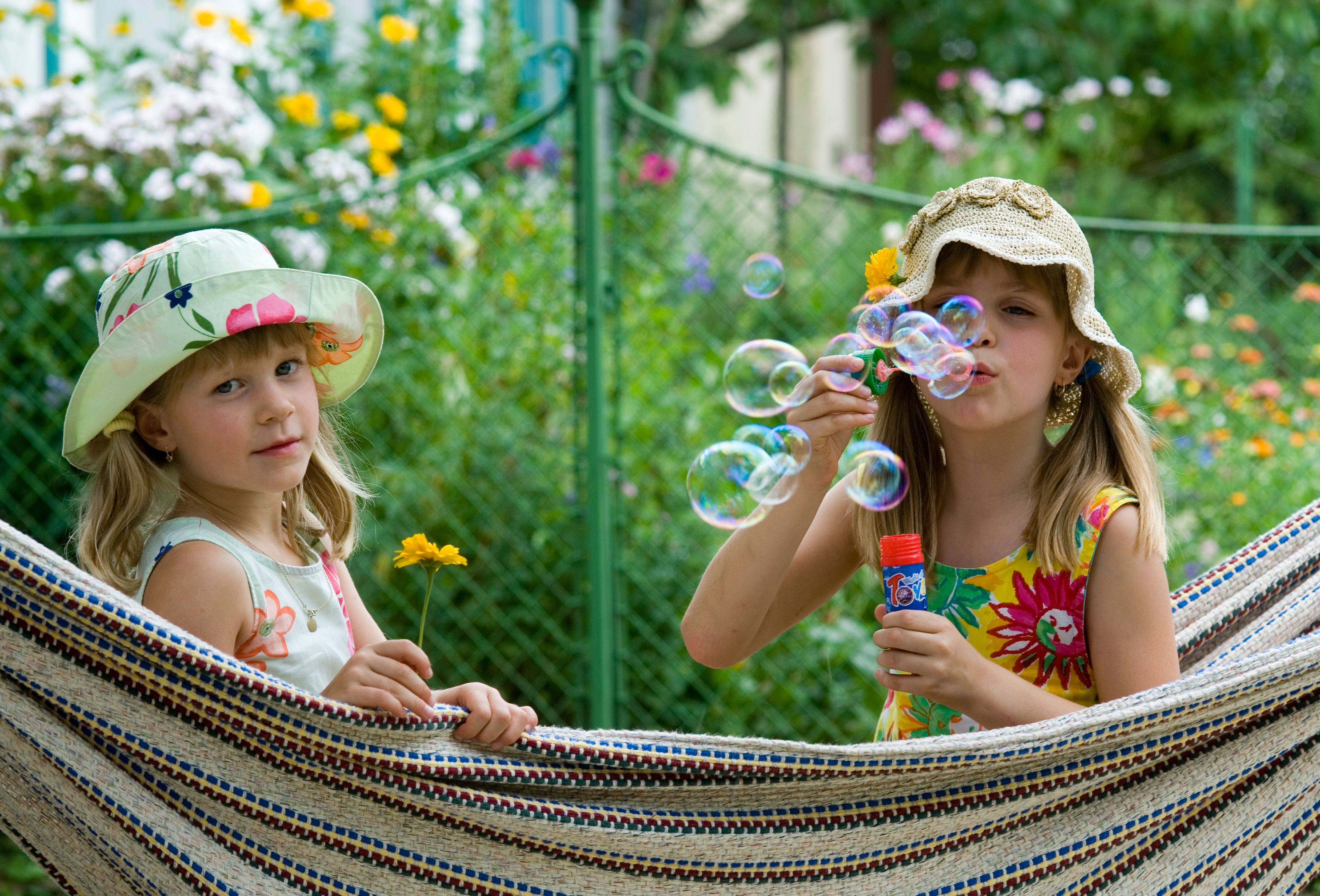 Two girls in summer dresses and hats are sitting in a hammock. One is blowing soap bubbles, the other is holding a yellow flower. Flowers blooming in the background.