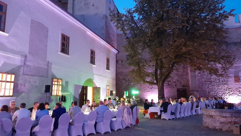 Abendveranstaltung im Freien mit gedeckten Tischen und Gästen vor einer beleuchteten historischen Mauer.