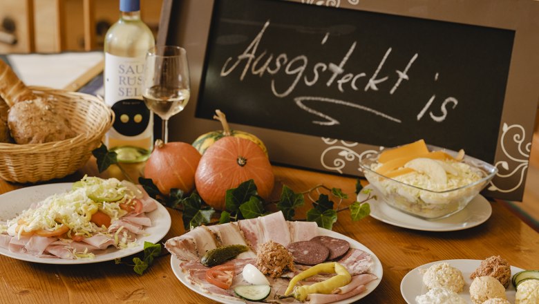 Ein gedeckter Tisch mit kalten Platten, Brot, Wein und einem Schild mit der Aufschrift 'Ausg'steckt is'.