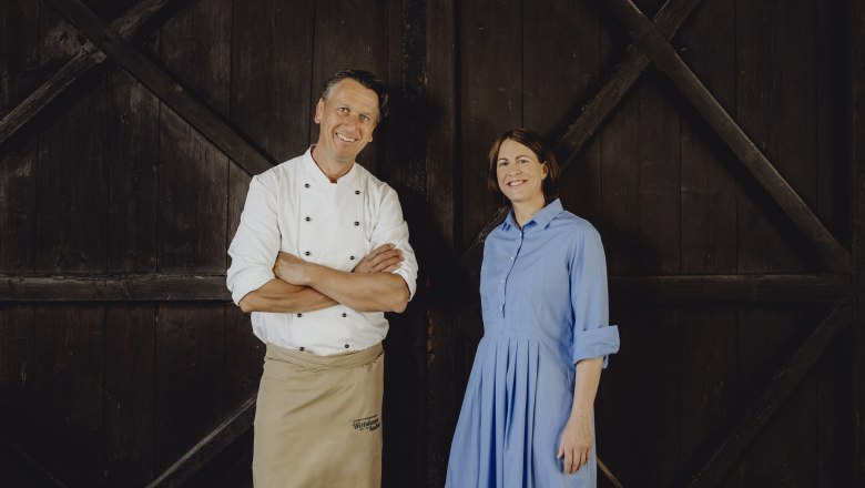 A man in chef's clothes and a woman in a blue dress stand in front of a wooden wall.