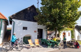 Menschen sitzen auf B&auml;nken vor einem wei&szlig;en Geb&auml;ude mit Fahrr&auml;dern in Alberndorf.