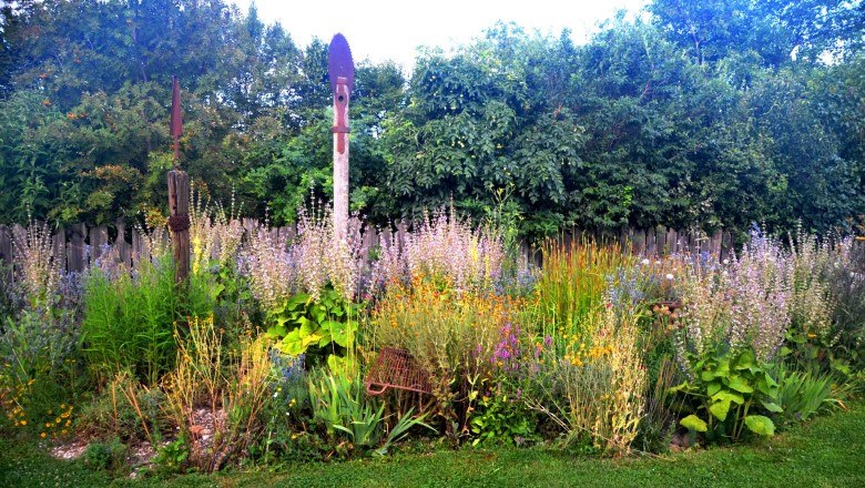 Ein buntes Staudenbeet mit verschiedenen Blumen und Pflanzen vor einem Holzzaun und Bäumen.