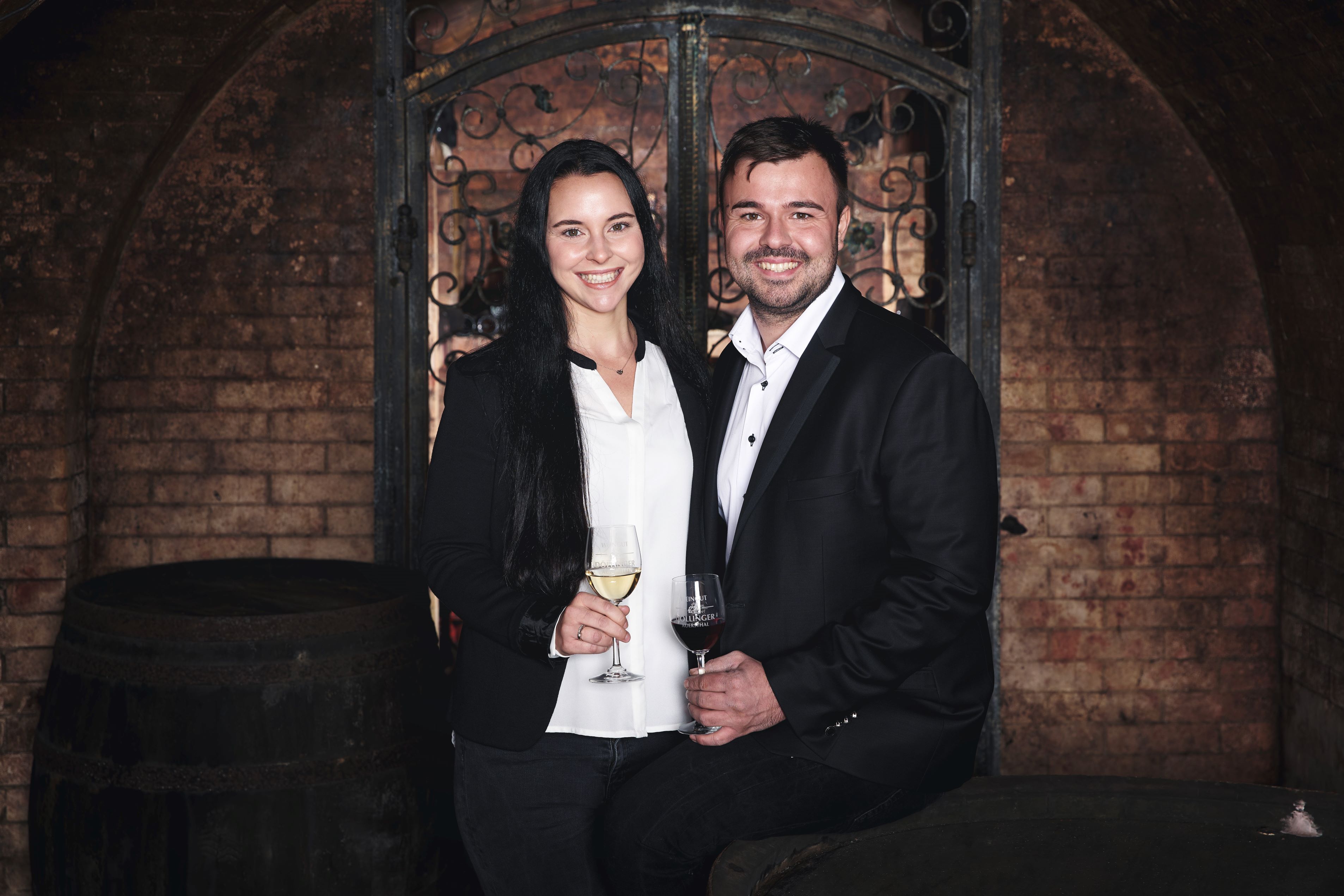 A man and a woman in elegant clothing hold wine glasses in a wine cellar.