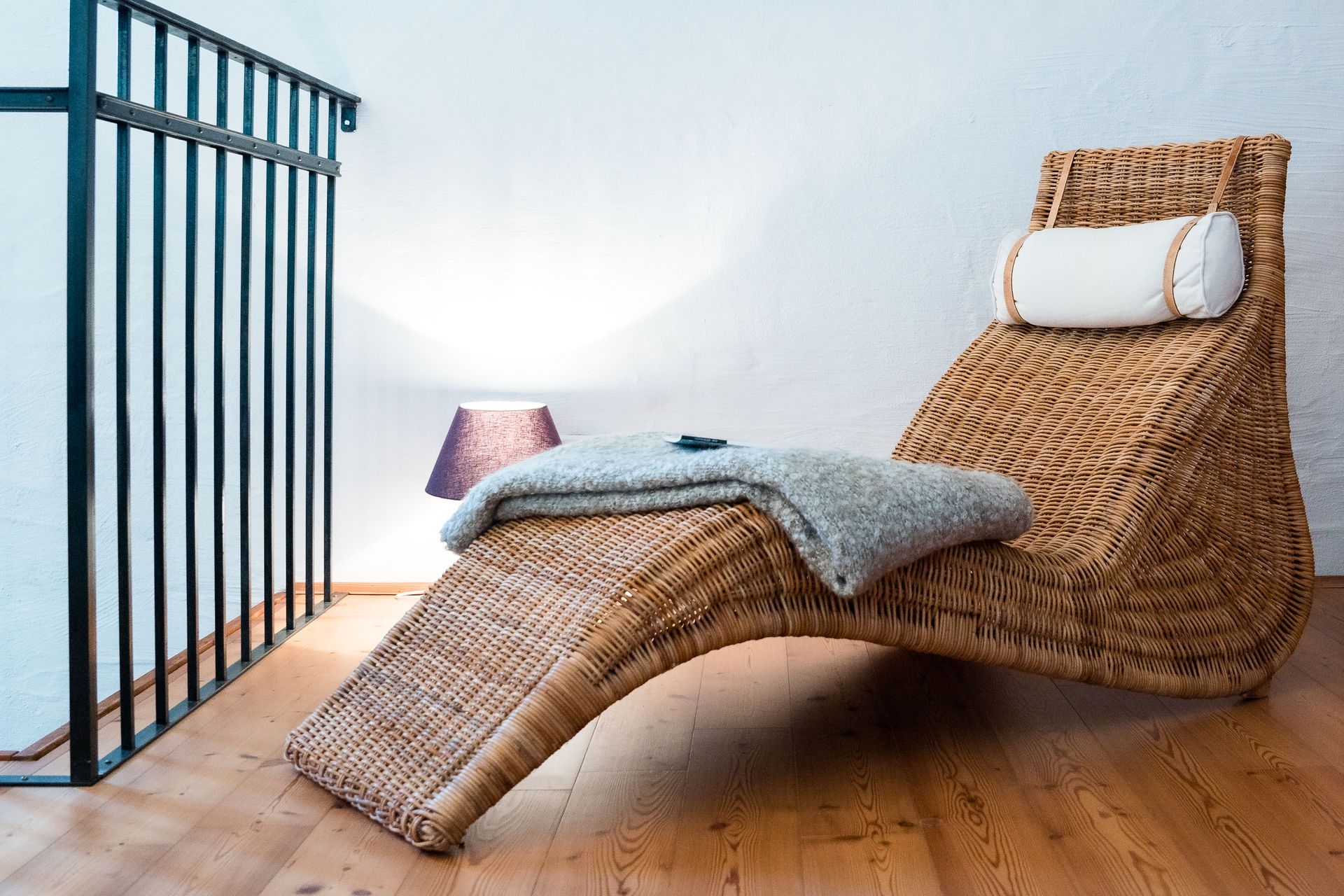 Eine gemütliche Leseecke mit einem geflochtenen Liegestuhl, einer Decke und einer Lampe auf Holzboden.