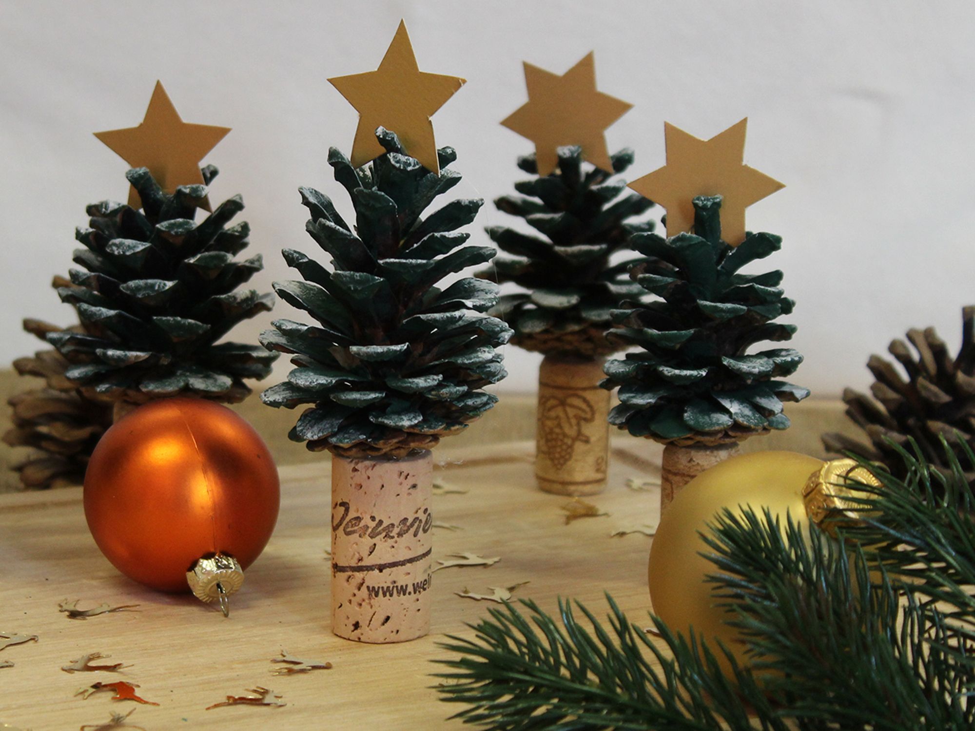 Mehrere Tannenbäumchen aus Korken und Zapfen stehen auf einem Holzbrett, rundherum liegen Weihnachtskugeln und Reisig. 