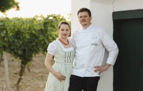 Ein Paar in traditioneller Kleidung steht vor einem weißen Gebäude neben einem Weinberg.