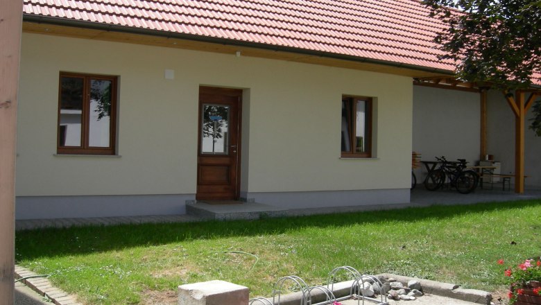 Ein Innenhof mit einem Gebäude mit rotem Ziegeldach, Holztür und Fenstern. Im Vordergrund sind Blumen und ein Fahrradständer zu sehen.