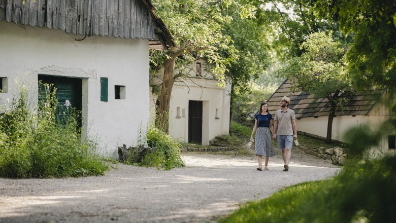 Ein Paar spaziert auf einem Weg zwischen alten Geb&auml;uden in einer gr&uuml;nen, l&auml;ndlichen Umgebung.
