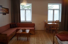 Gemütliches Zimmer mit Holzmöbeln, Sofa, Tisch und Stühlen, zwei Fenstern und einem Bild an der Wand.