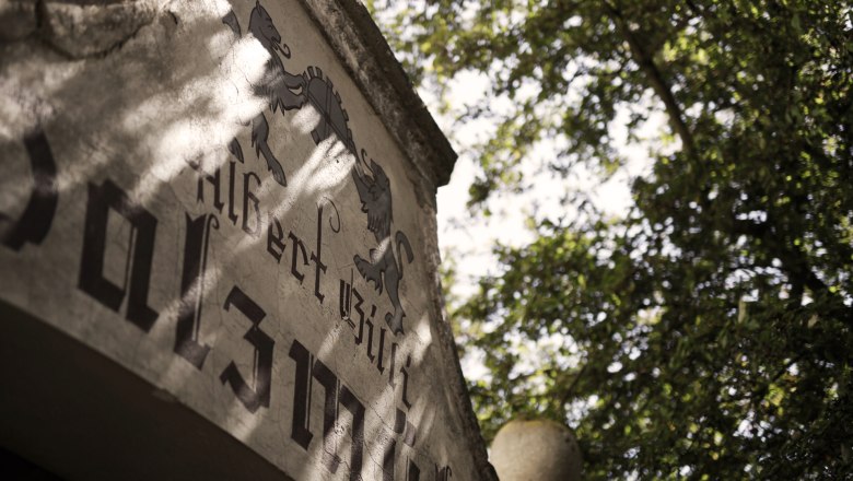 Close-up of an old building with lettering and coat of arms, surrounded by trees.