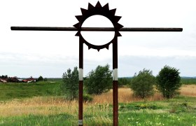 Metallskulptur in Form einer Sonne auf einer Wiese mit Bäumen im Hintergrund.