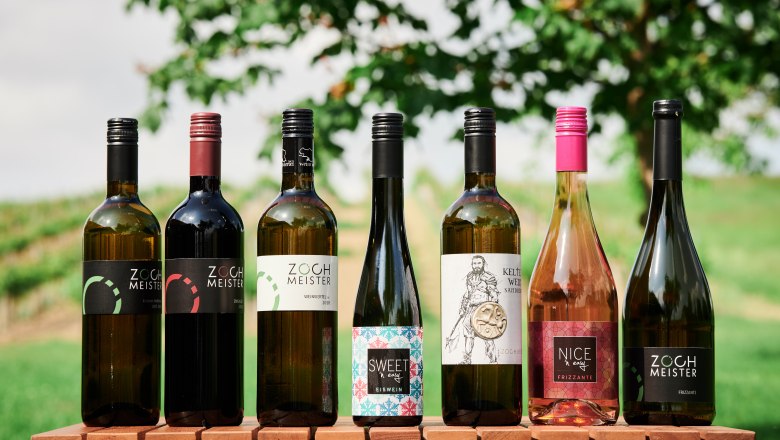 Sieben Weinflaschen auf einem Holztisch vor einem unscharfen Hintergrund mit B&auml;umen und Wiese.