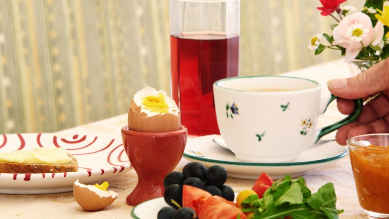 Frühstückstisch mit gekochtem Ei, Saft, Kaffee, Brot, Obst und Blumen.