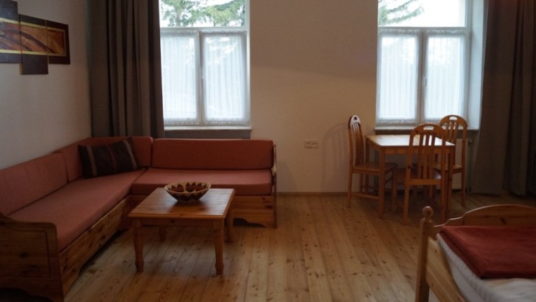 Zimmeransicht, © © Meisel Gemütliches Zimmer mit Holzmöbeln, Sofa, Tisch und Stühlen, zwei Fenstern und einem Bild an der Wand.