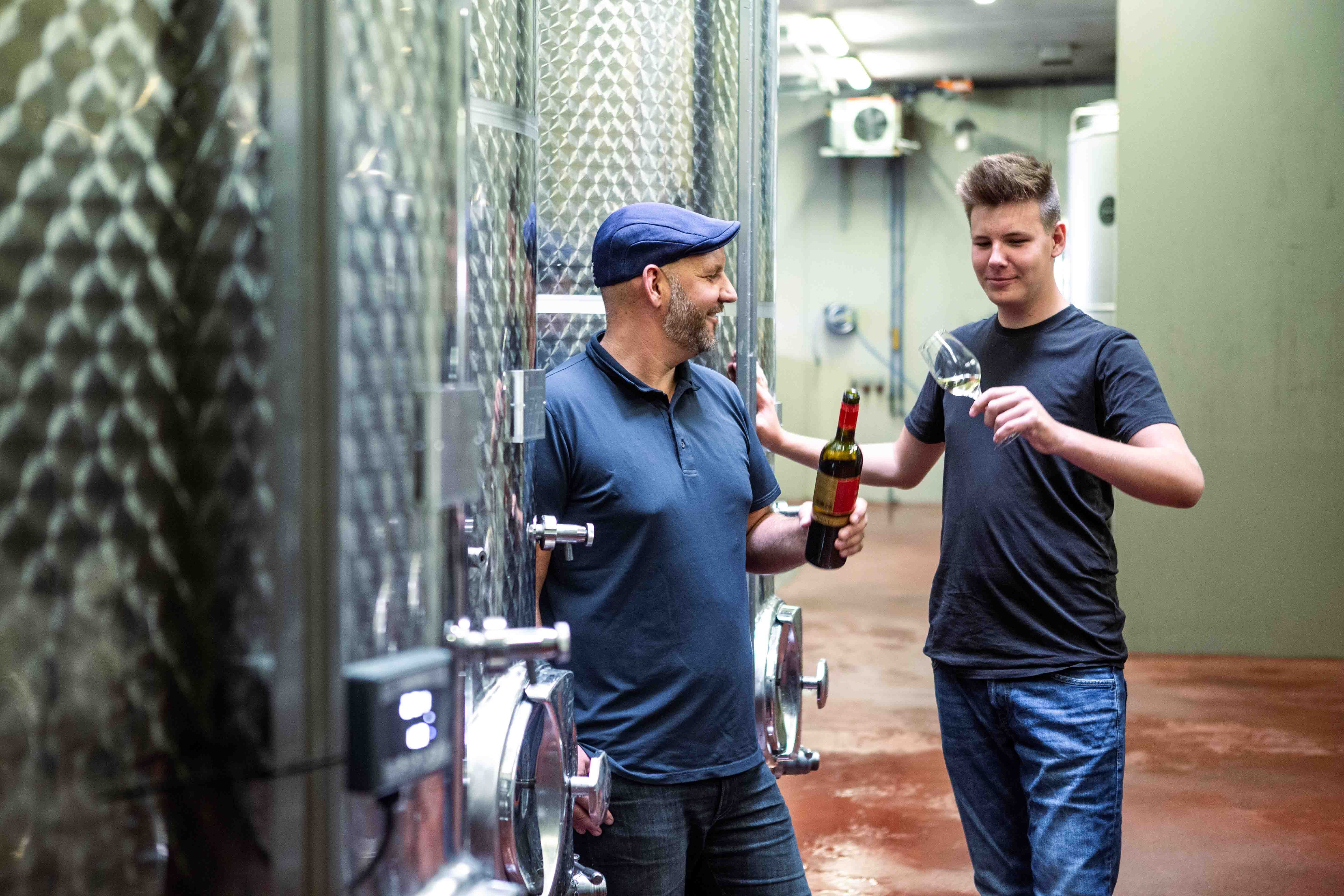 Two men in a wine cellar, one holding a wine bottle, the other a wine glass.