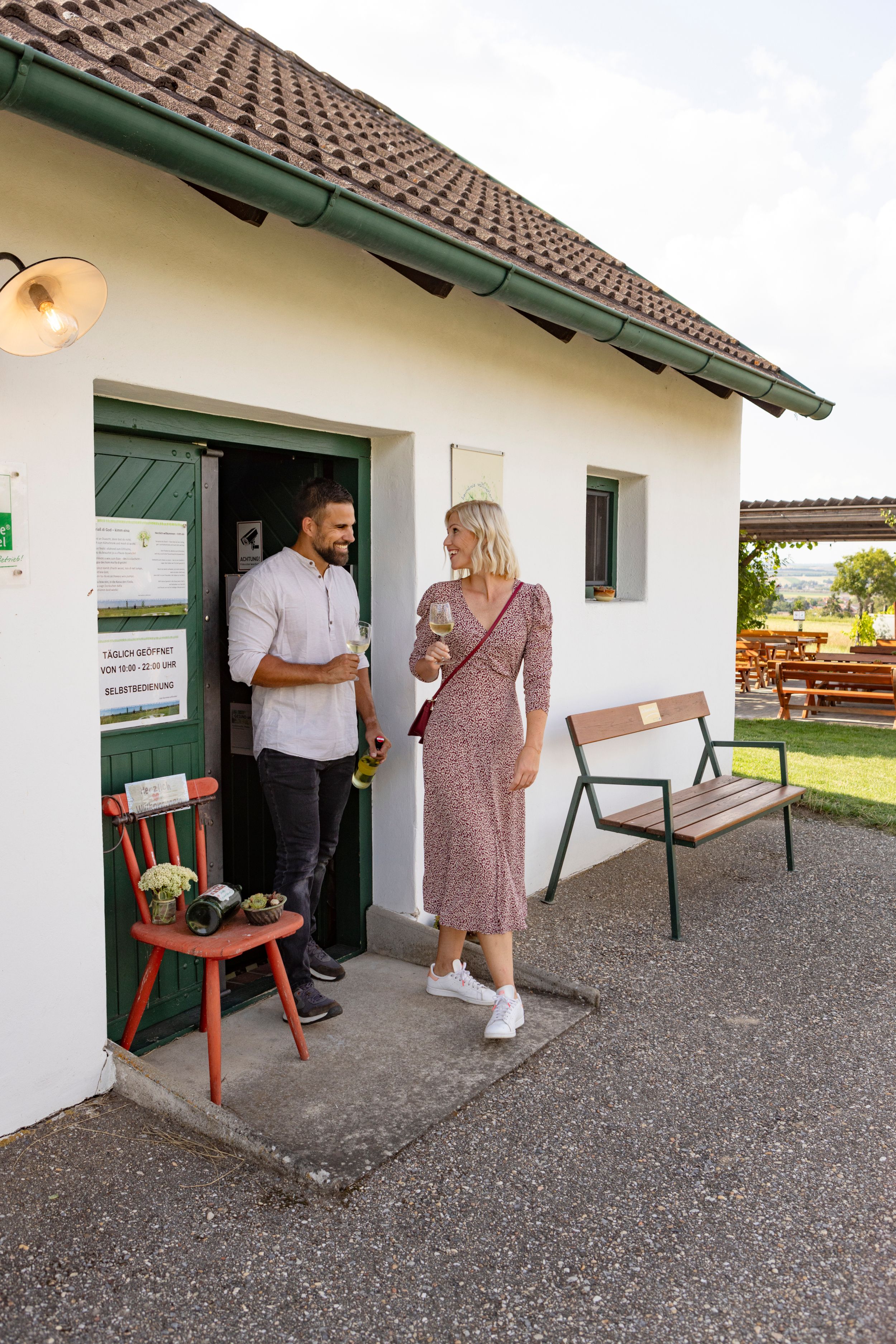 A man and a woman are standing in front of a small white building drinking wine.
