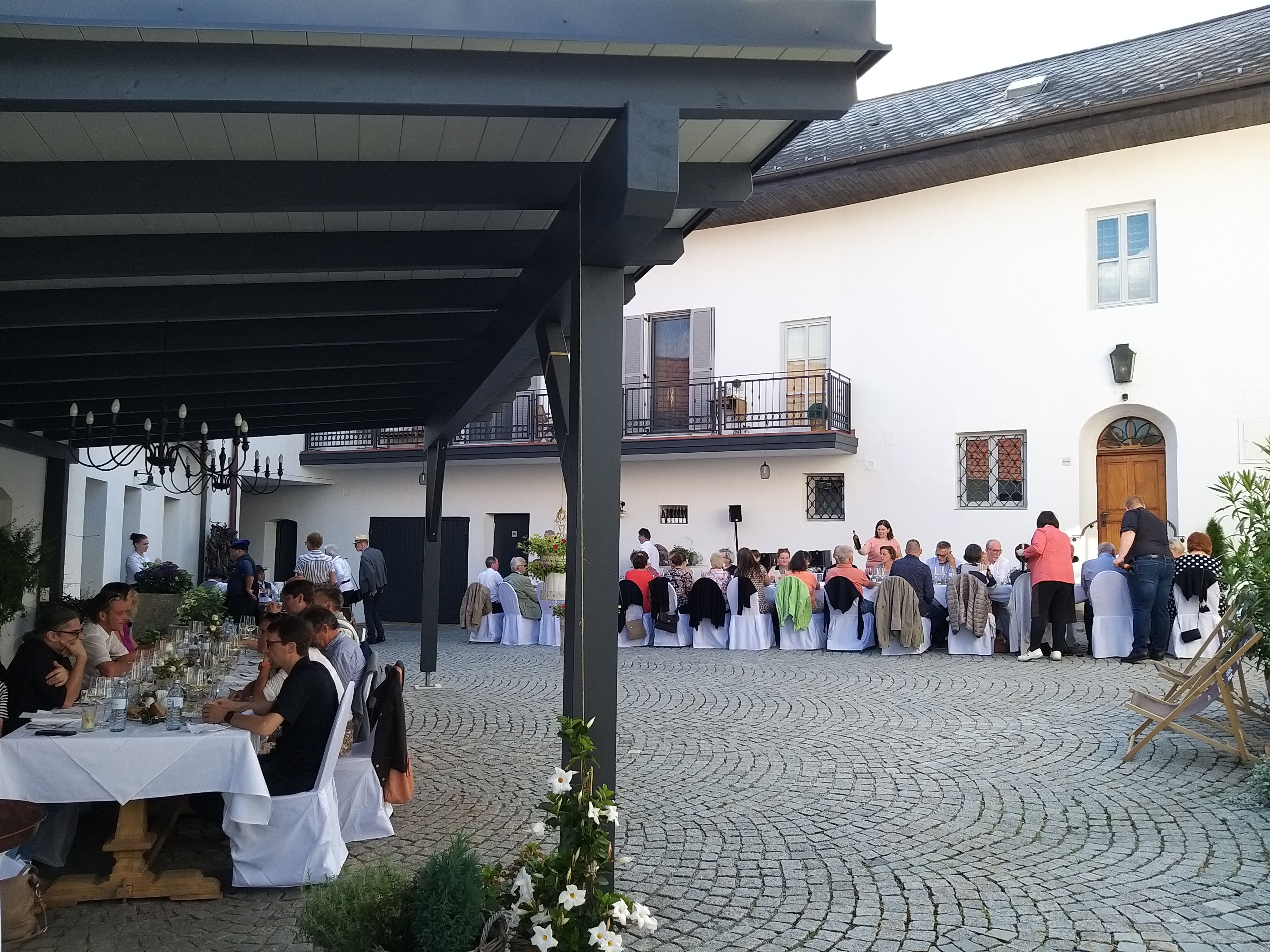 Menschen sitzen an langen Tischen in einem gepflasterten Innenhof und essen.