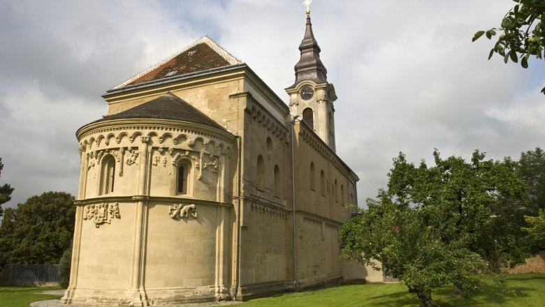 Romanische Kirche mit Verzierungen und Turm, umgeben von grüner Wiese und Bäumen.