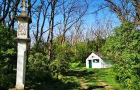 Ein steinernes Kreuz und ein kleines wei&szlig;es Geb&auml;ude in einem Waldgebiet bei sonnigem Wetter.