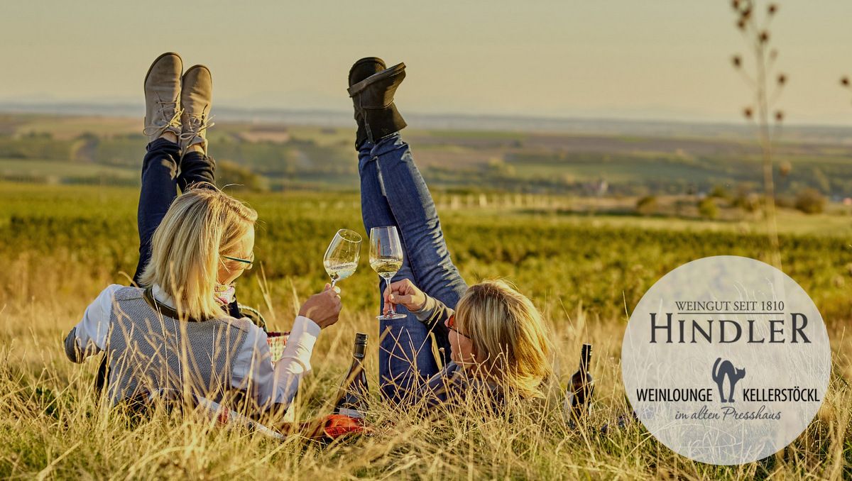 Zwei Personen liegen in einem Weinberg, heben die Füße und halten Weingläser. Logo des Weinguts Hindler ist eingeblendet.