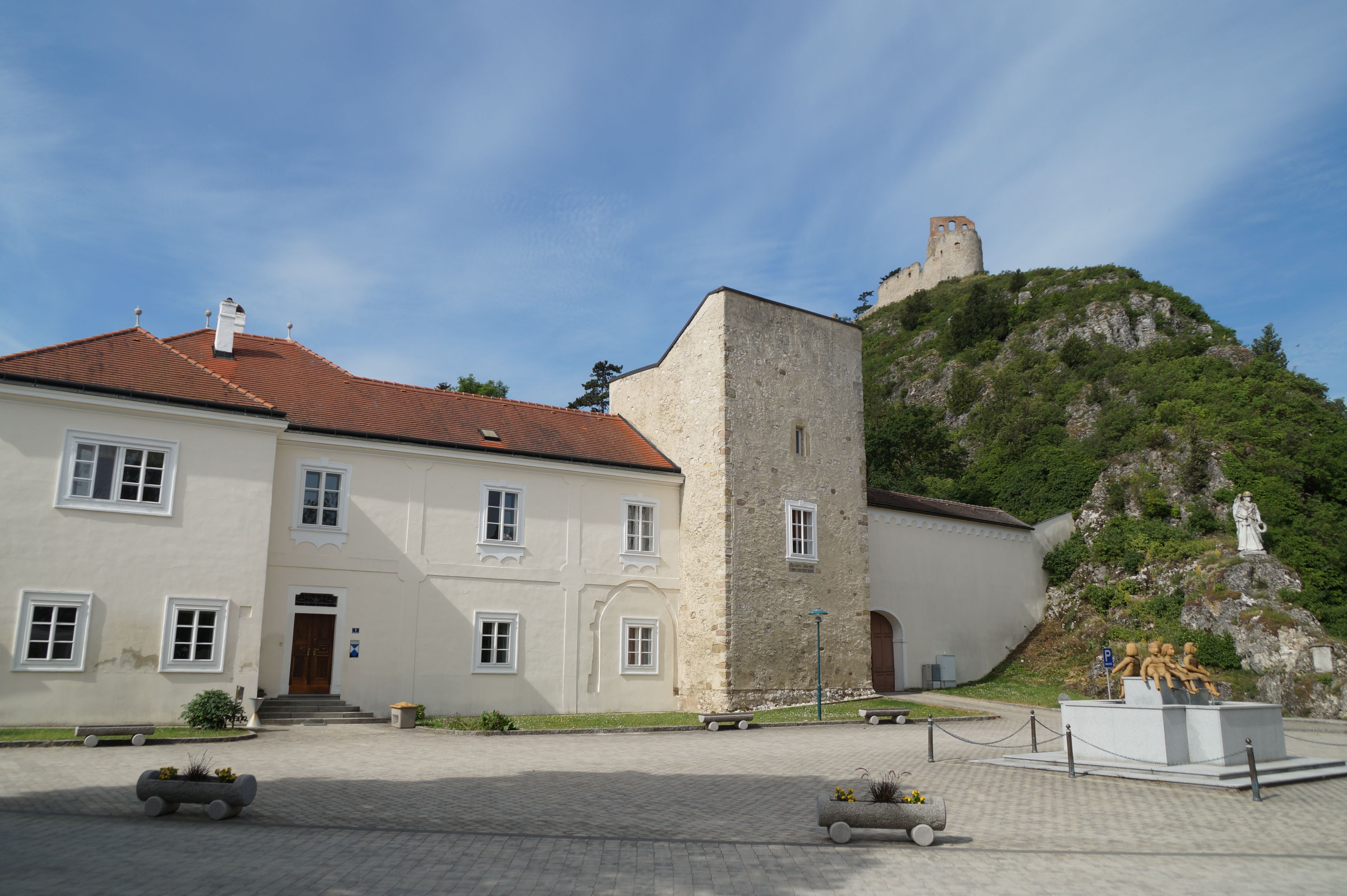 Historická budova s červenou taškovou střechou a kamennou věží, v pozadí zřícenina hradu na kopci.