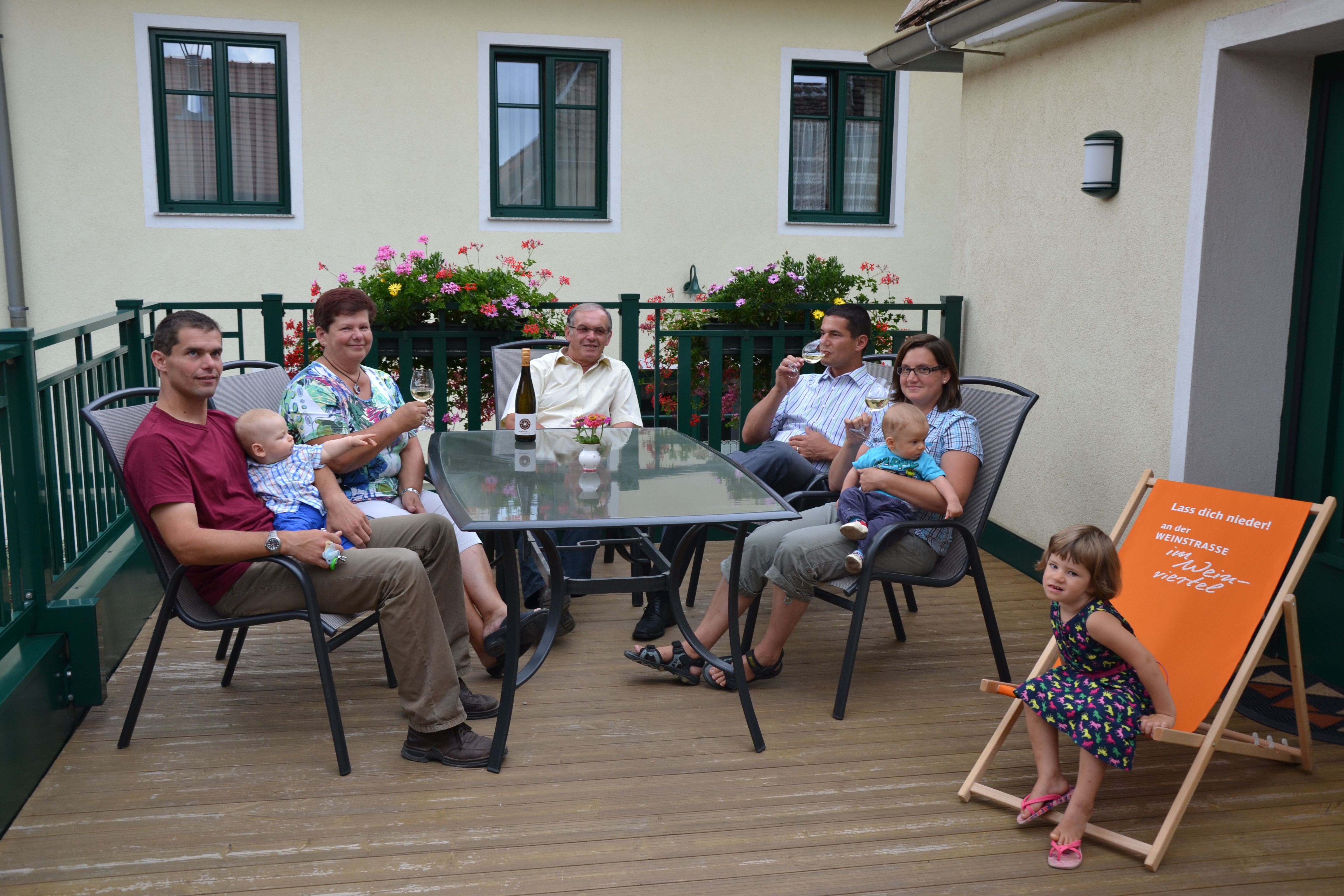 Eine Familie sitzt auf einer Terrasse an einem Tisch mit Getränken. Im Hintergrund sind Blumen und ein Gebäude zu sehen.