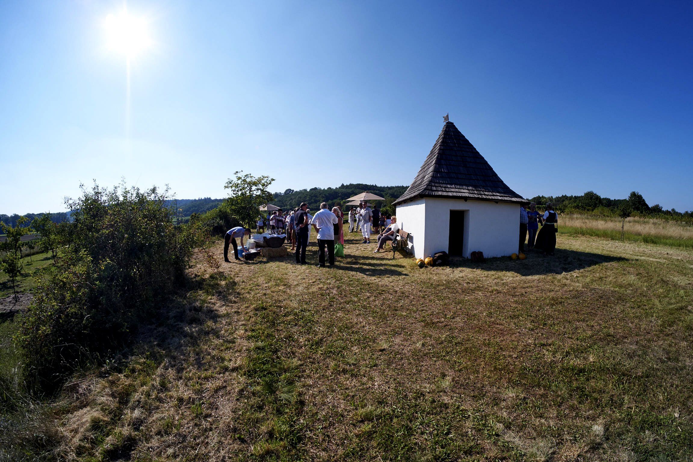 Eine Gruppe von Menschen versammelt sich bei einer kleinen, weißen Hütte mit spitzem Dach auf einer Wiese unter klarem Himmel.