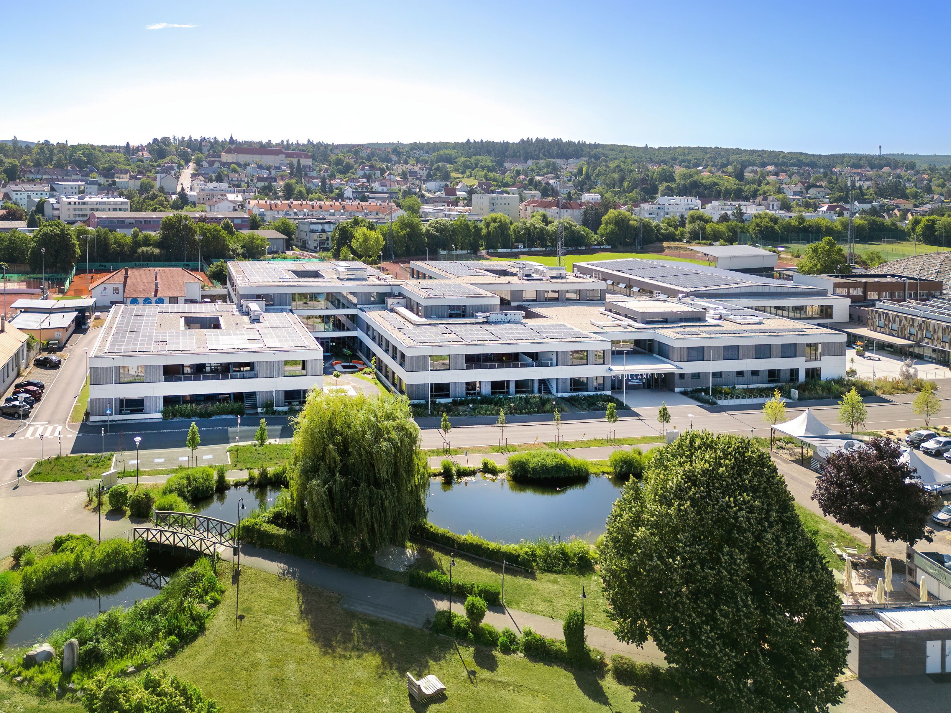Luftaufnahme eines modernen Schulcampus mit Solarpanelen auf den Dächern, umgeben von Grünflächen und einem Teich im Vordergrund.
