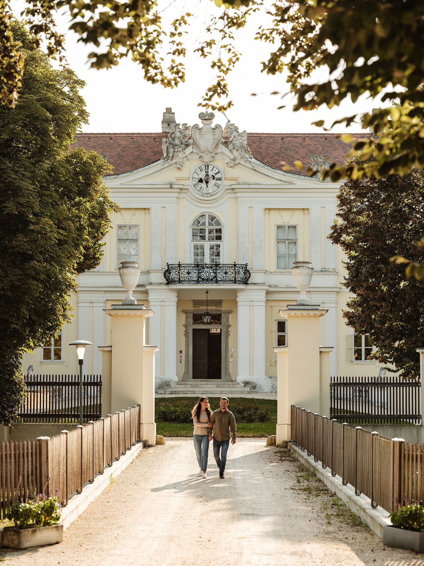 Ein malerischer Weg führt zu einem prächtigen Schloss, umgeben von üppigem Grün und majestätischen Bäumen. Die sanfte Abendsonne taucht die Fassade in warmes Licht und schafft eine einladende Atmosphäre für Besucher, die die Schönheit der Umgebung genießen möchten.