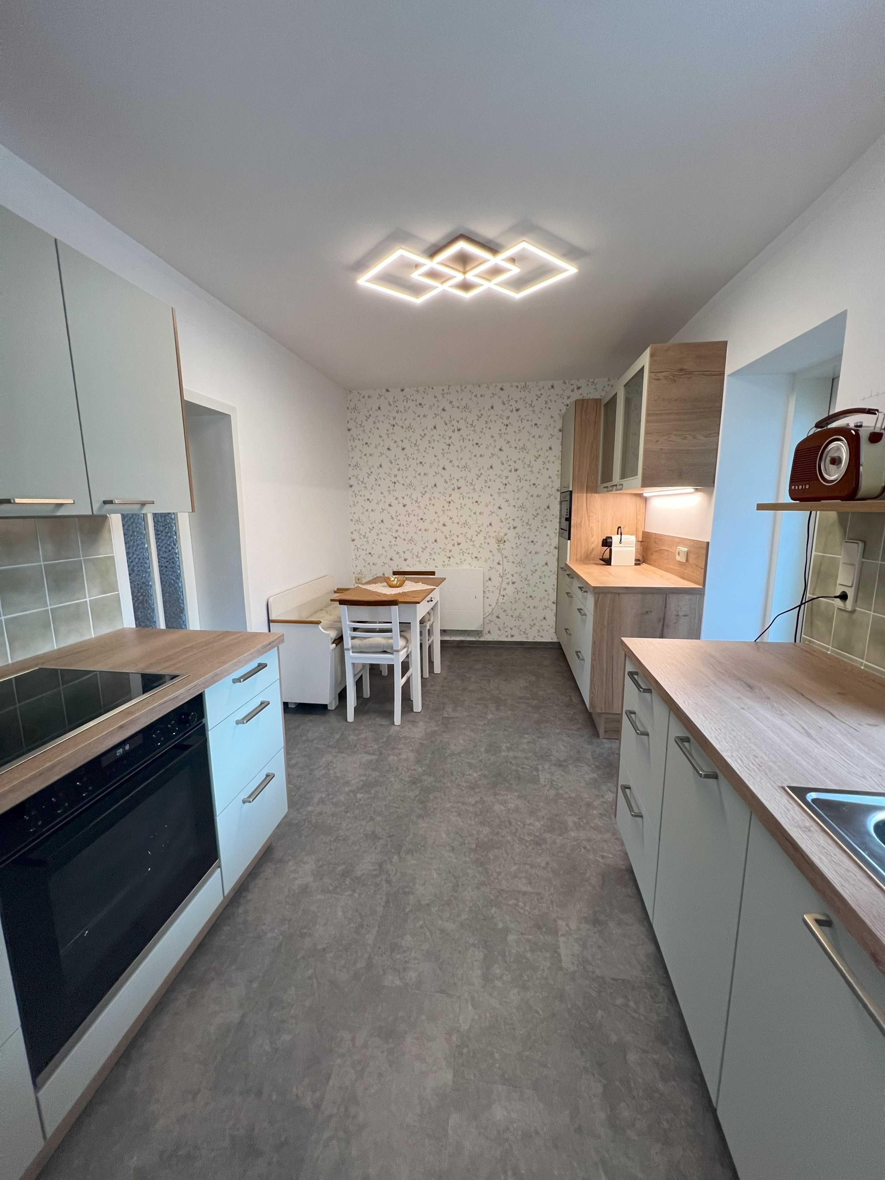 Moderne Küche mit grauen Schränken, Holzakzenten und einem Esstisch. An der Decke eine geometrische Lampe.