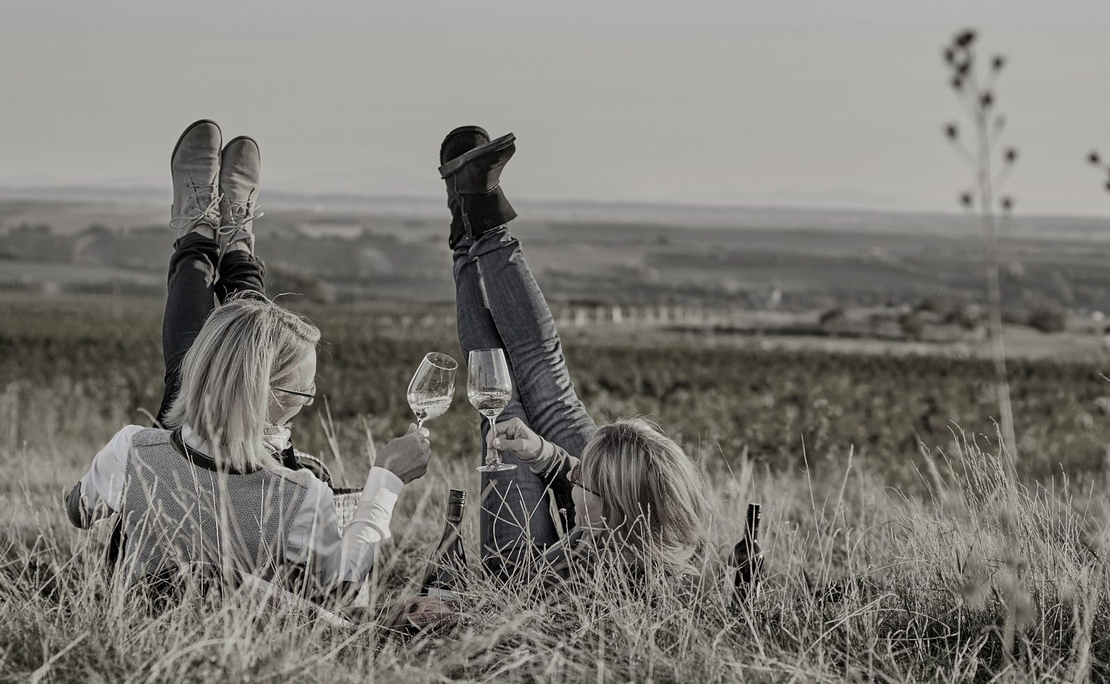 Zwei Personen liegen im Gras mit Weingläsern in der Hand, die Beine in die Luft gestreckt, vor einer weiten Landschaft.