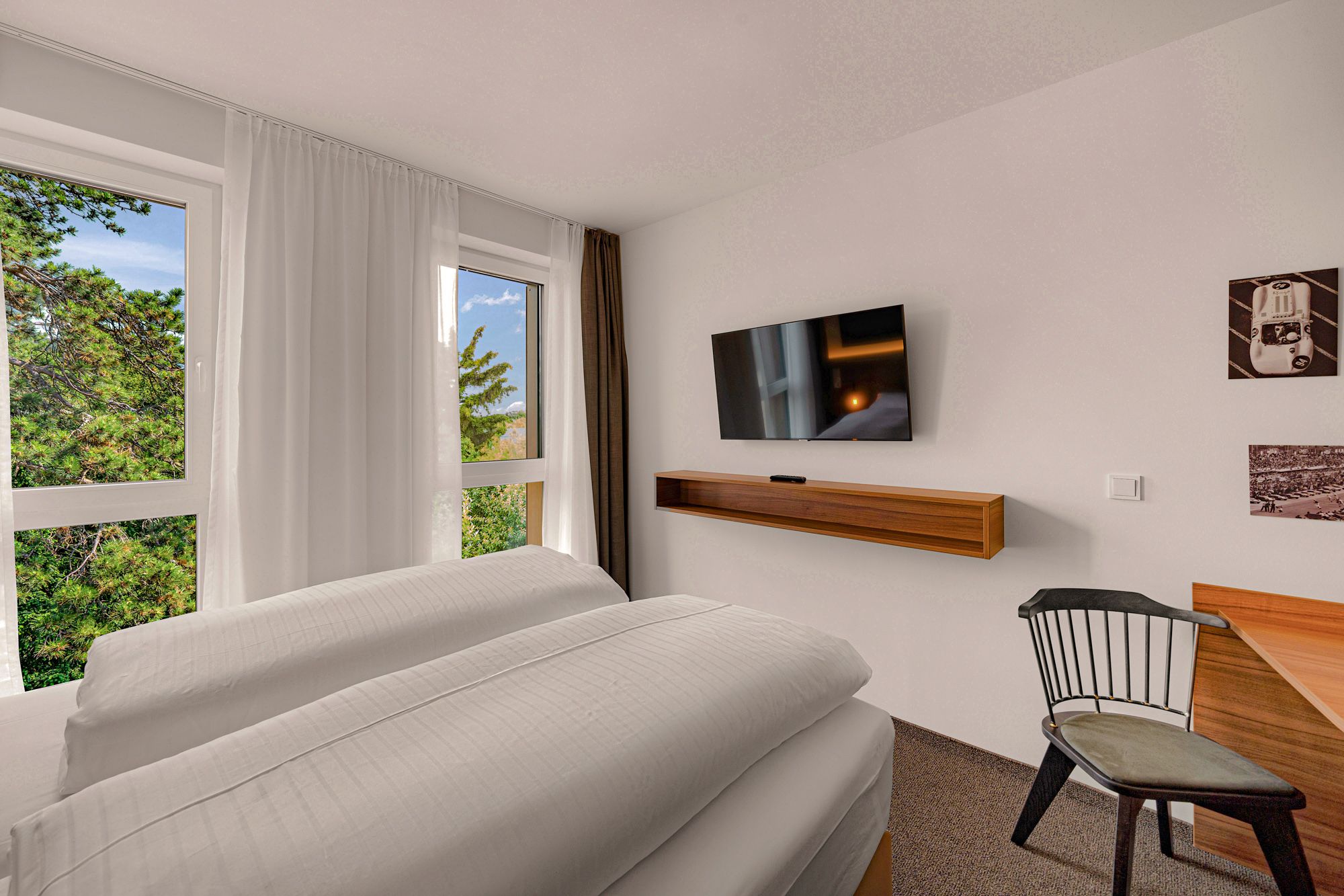 Modernes Hotelzimmer mit großem Bett, Flachbildfernseher, Schreibtisch und Stuhl. Fenster mit Blick auf Bäume.