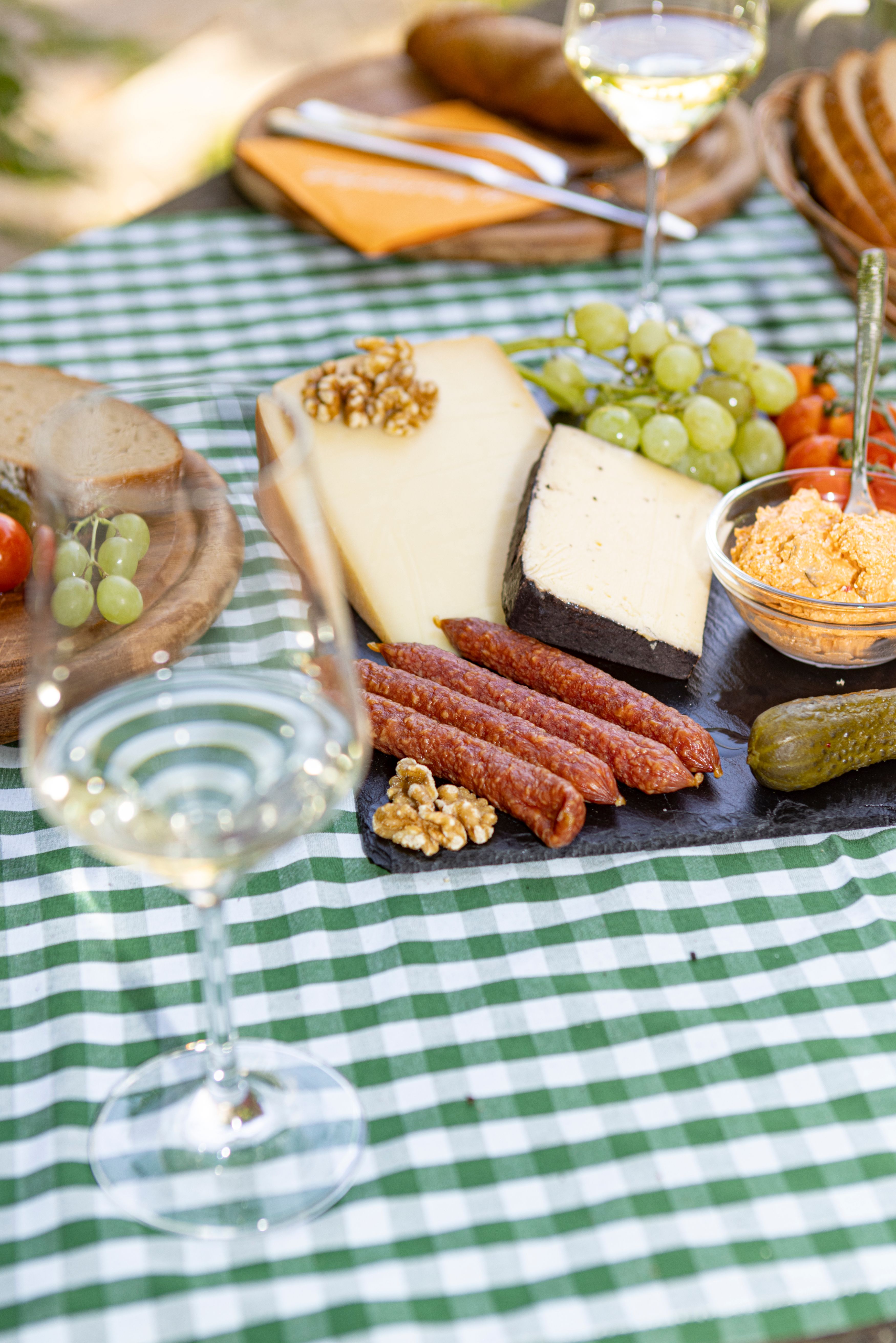 Picnic table with cheese, sausage, bread, grapes and a glass of white wine on a checkered tablecloth.