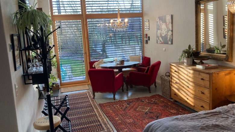 Wohnzimmer mit großen Fenstern, roten Sesseln, Holzmöbeln und Teppichen, Blick in den Garten.