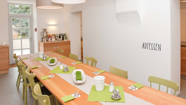 Ein heller Speiseraum mit einem langen Holztisch, grünen Stühlen und Dekoration. An der Wand steht 'AUFESSEN'.