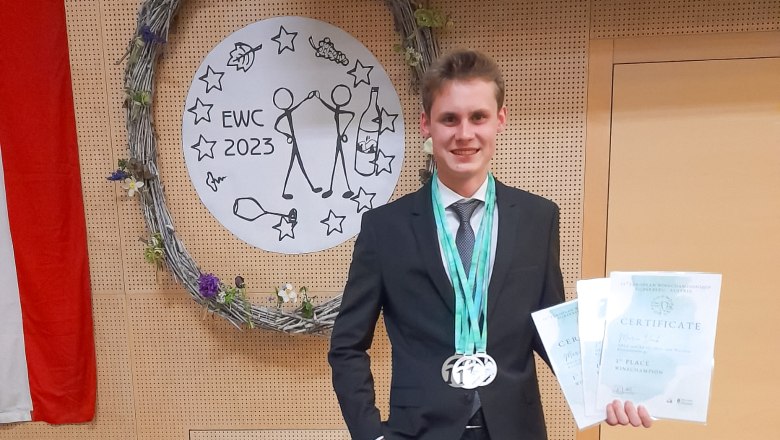 Ein junger Mann im Anzug steht vor einem dekorativen Kranz mit einem Zertifikat und Medaillen in der Hand.