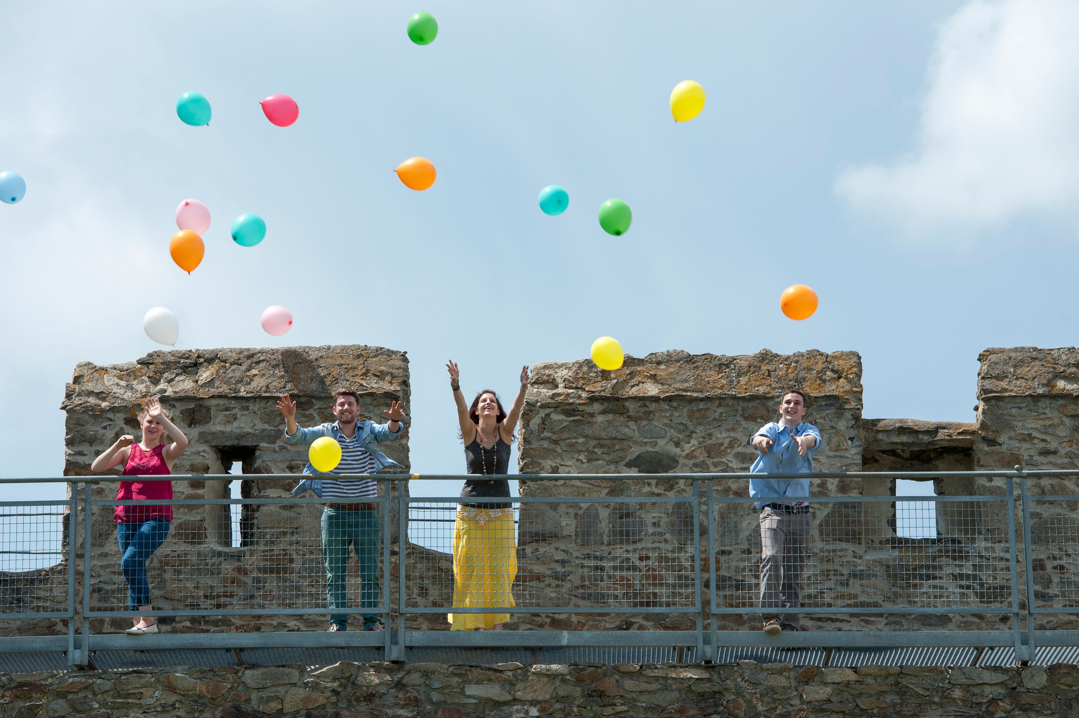 Four people throw colorful balloons into the air from a castle wall.
