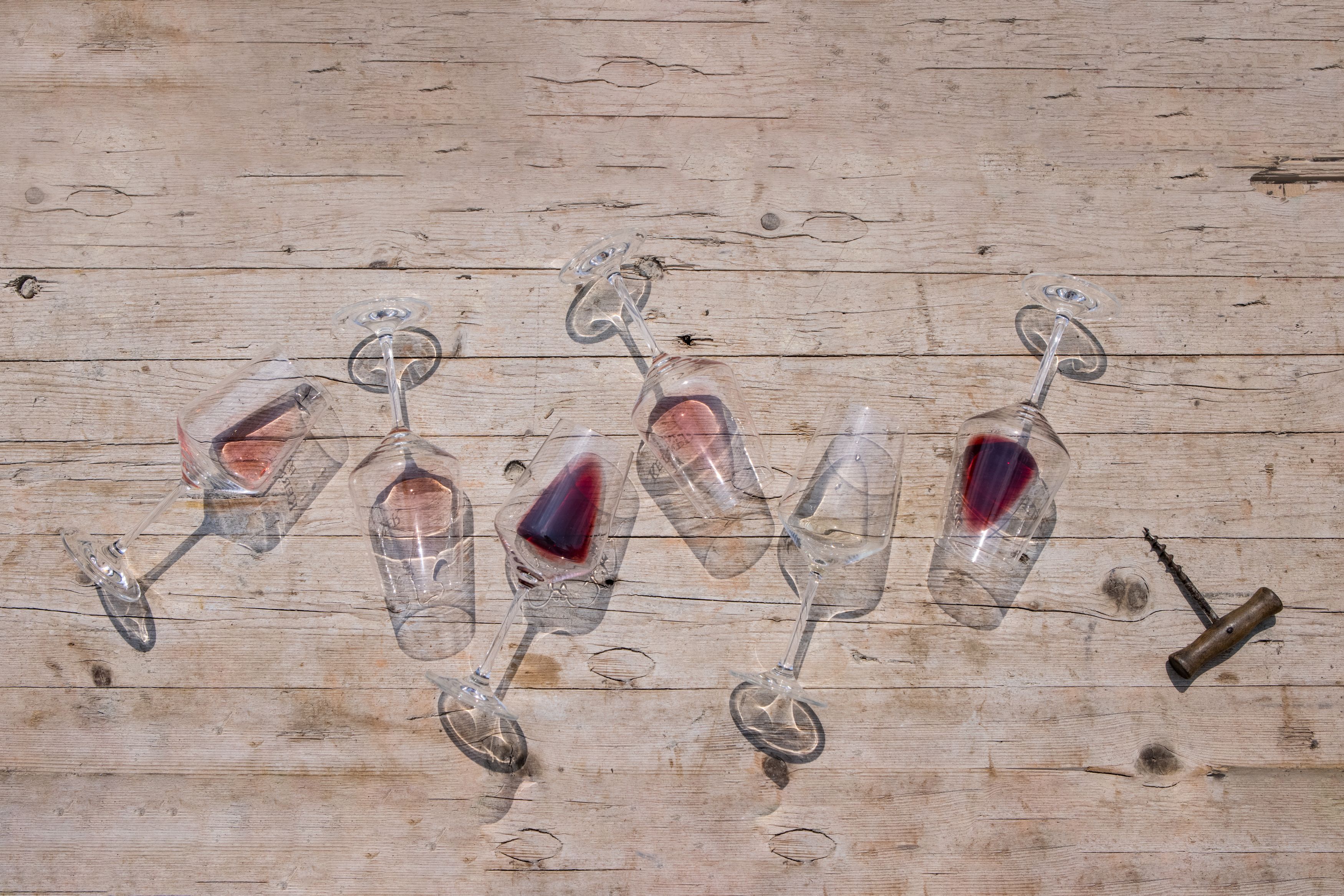 Six overturned wine glasses with red wine on a wooden table, a corkscrew next to them.