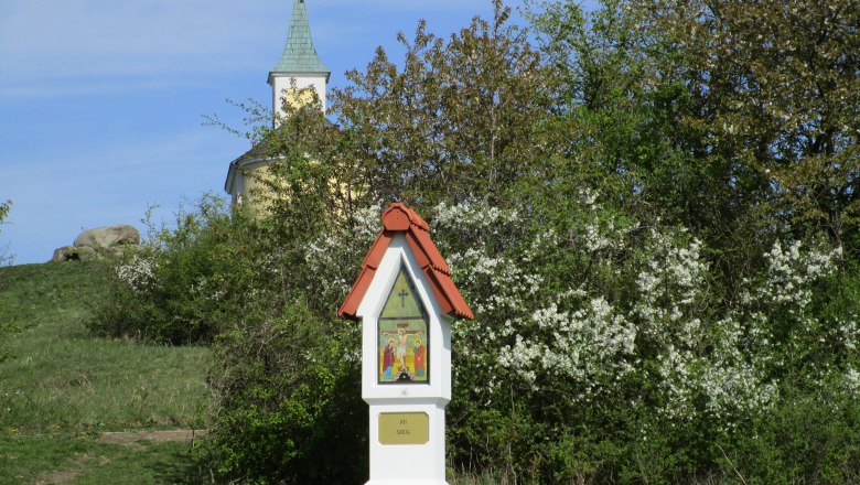 Eine kleine Kapelle mit grünem Turm hinter blühenden Büschen und einem Bildstock im Vordergrund.