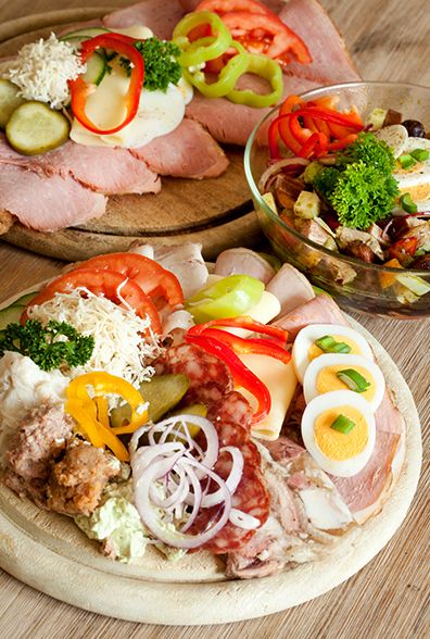 Various types of sausage and cheese on a wooden board, garnished with vegetables and eggs, next to a salad in a glass bowl.