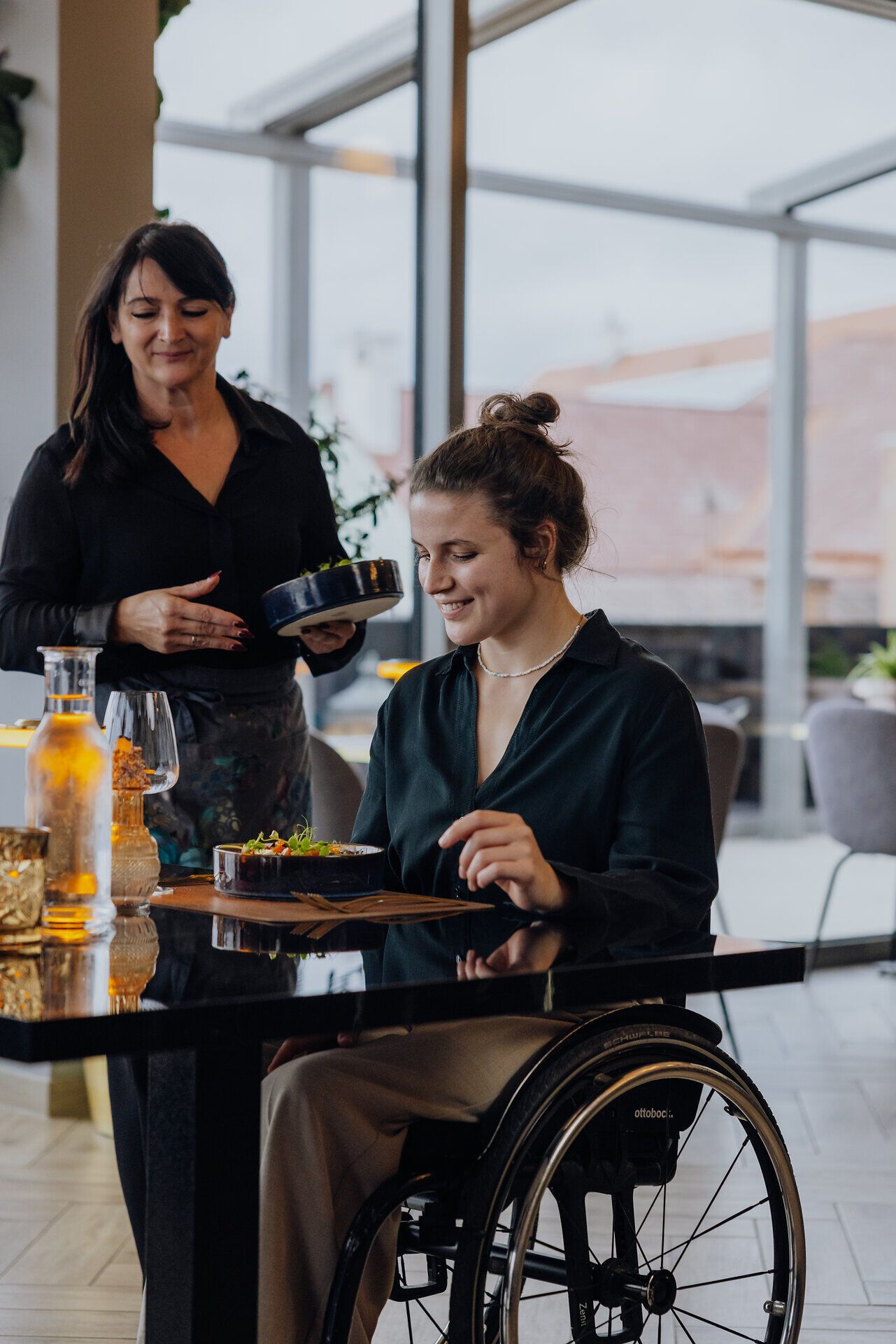 Eine junge Frau im Rollstuhl sitzt in einem Restaurant an einem Tisch und bekommt von einer Kellnerin eine Speise serviert. 