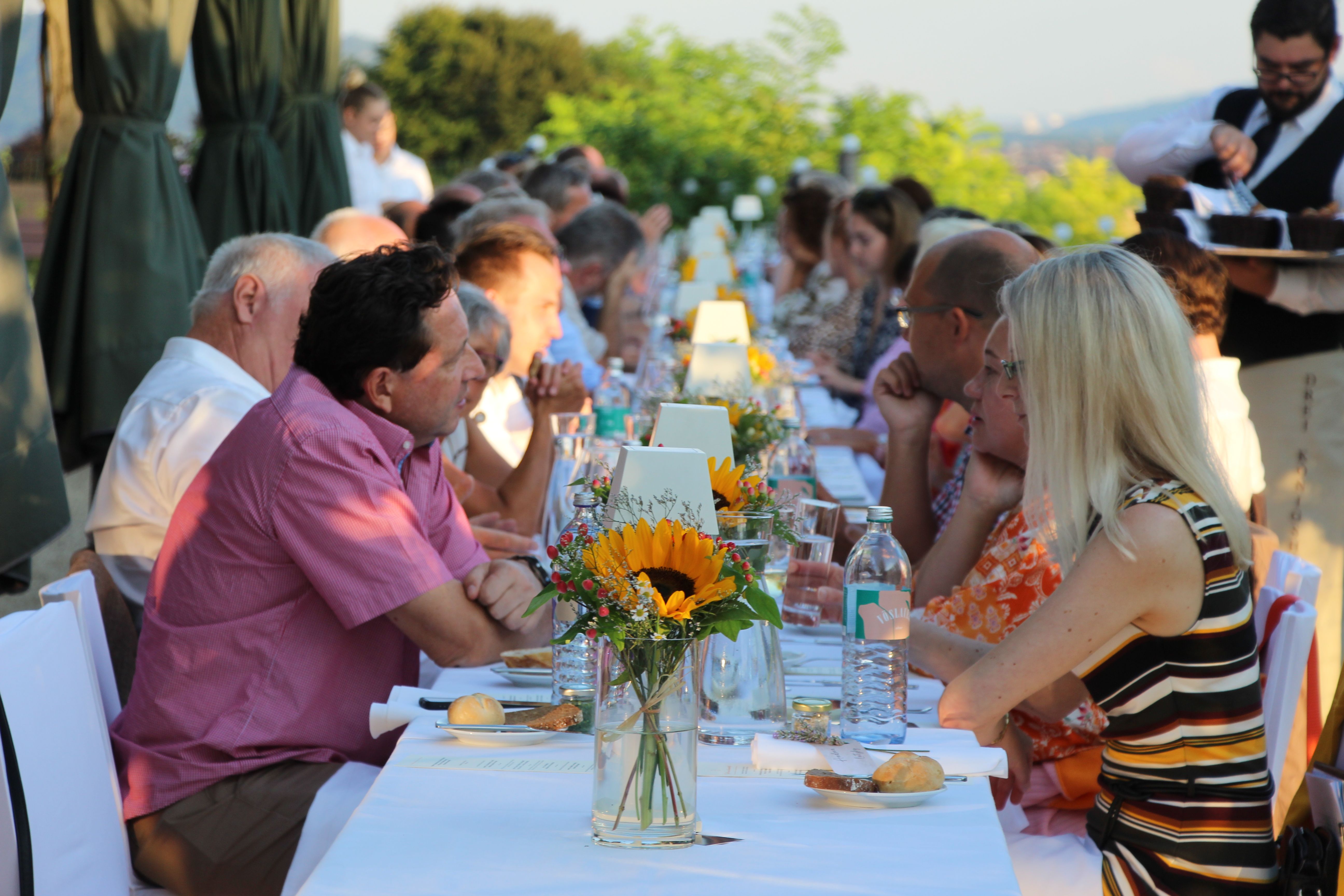 Menschen sitzen an einer langen, festlich gedeckten Tafel im Freien.