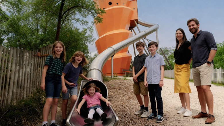 Kinder spielen auf einer Rutsche im Freien mit einem gro&szlig;en orangefarbenen Turm im Hintergrund.