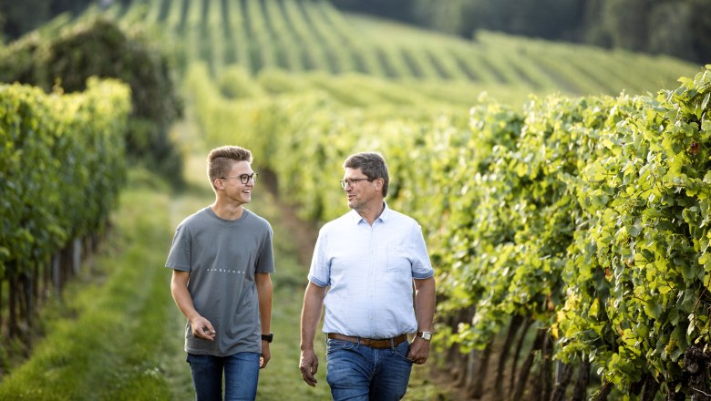 Two men walk through a vineyard smiling.