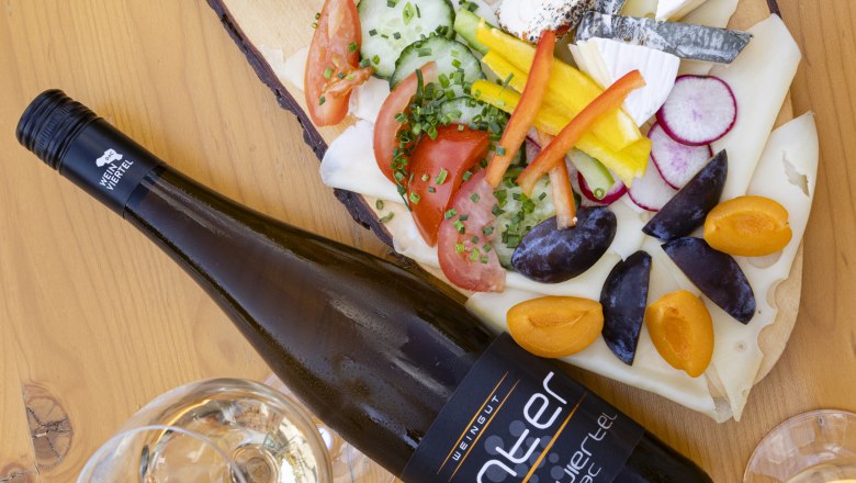 Eine Heurigenplatte mit verschiedenen Käsesorten, Aufstrichen, Gemüse und einer Flasche Wein auf einem Holztisch.
