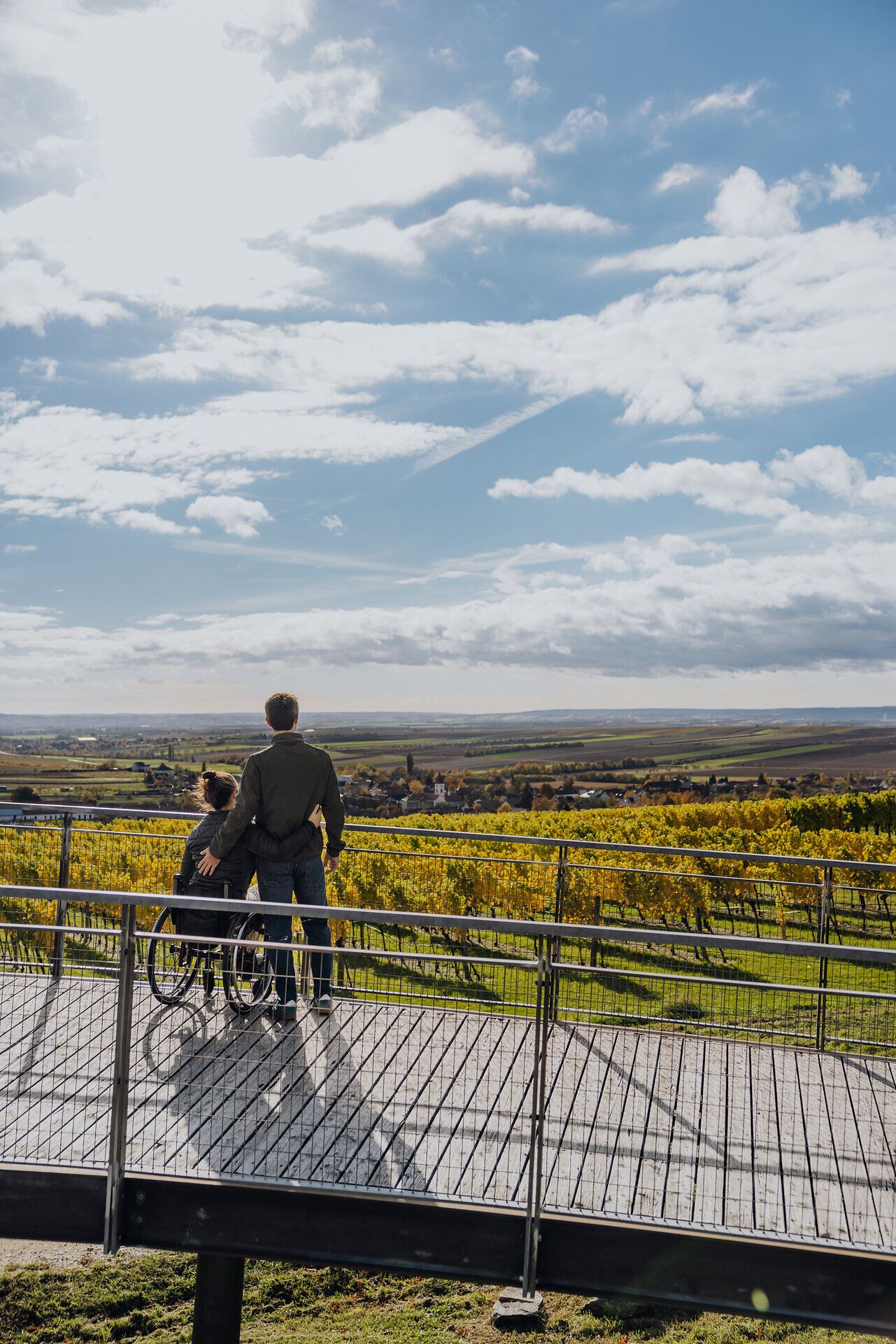 Eine Frau im Rollstuhl und ein Mann stehen auf einer Aussichtsplattform und blicken auf die herbstlich gefärbten Weingärten und die Weite der Weinviertler Landschaft.
