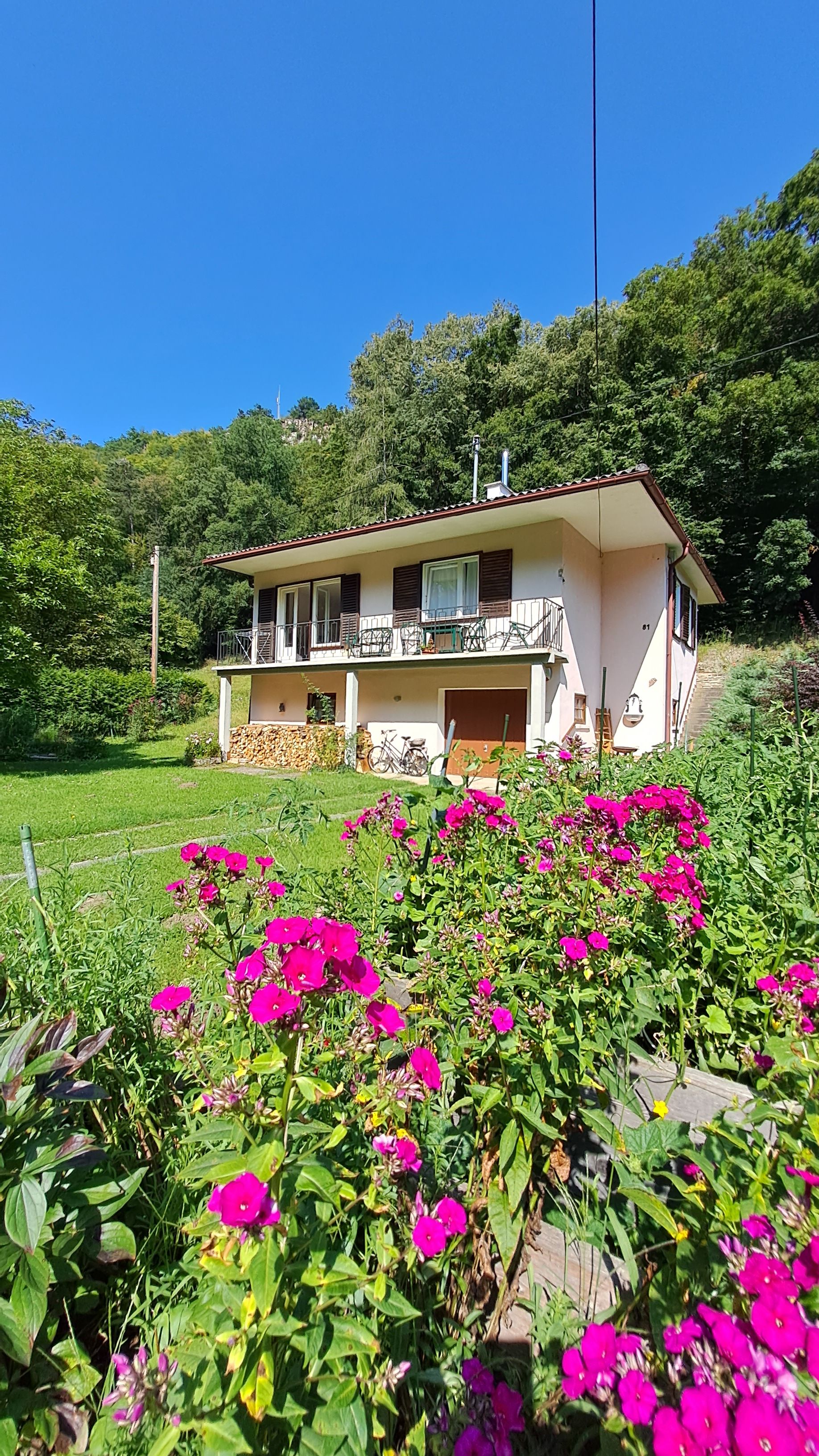 Ein weißes Haus mit Balkon und Holzstapel im Garten, umgeben von grünen Bäumen und pinken Blumen im Vordergrund.