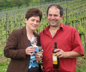 A man and a woman are standing in a vineyard holding wine glasses and a bottle of wine.
